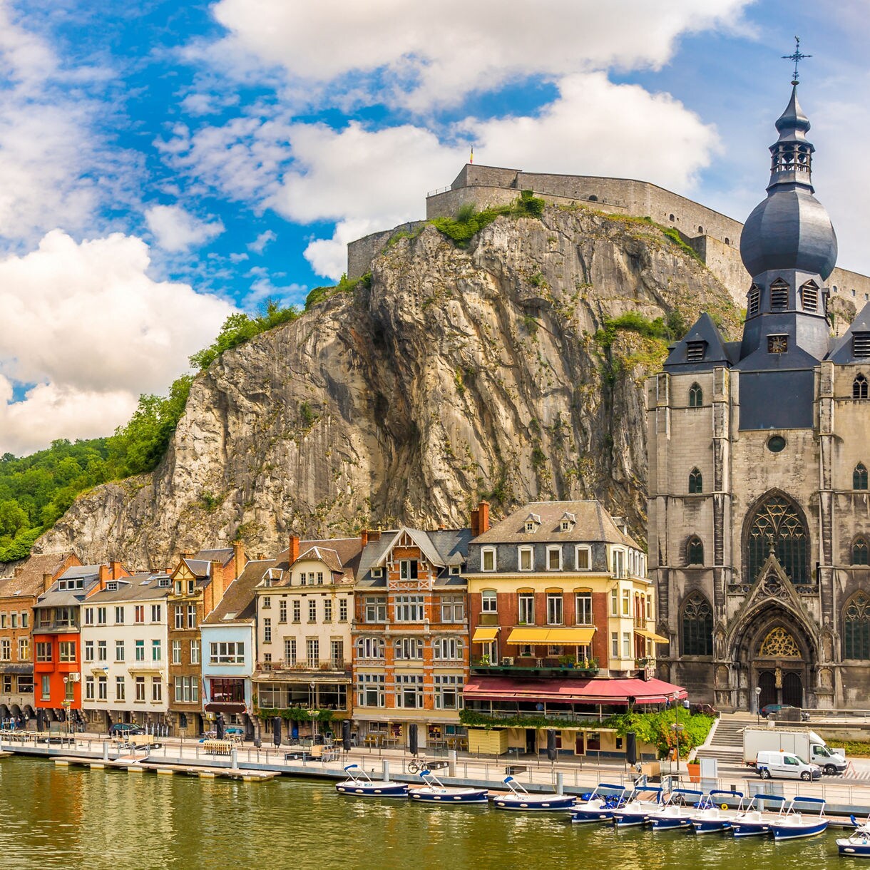 Panoramic view of Dinant, Belgium, showing the Meuse River with boats, rows of colorful buildings, the Collegiate Church of Our Lady with its onion dome and a fortress perched on the cliff above.