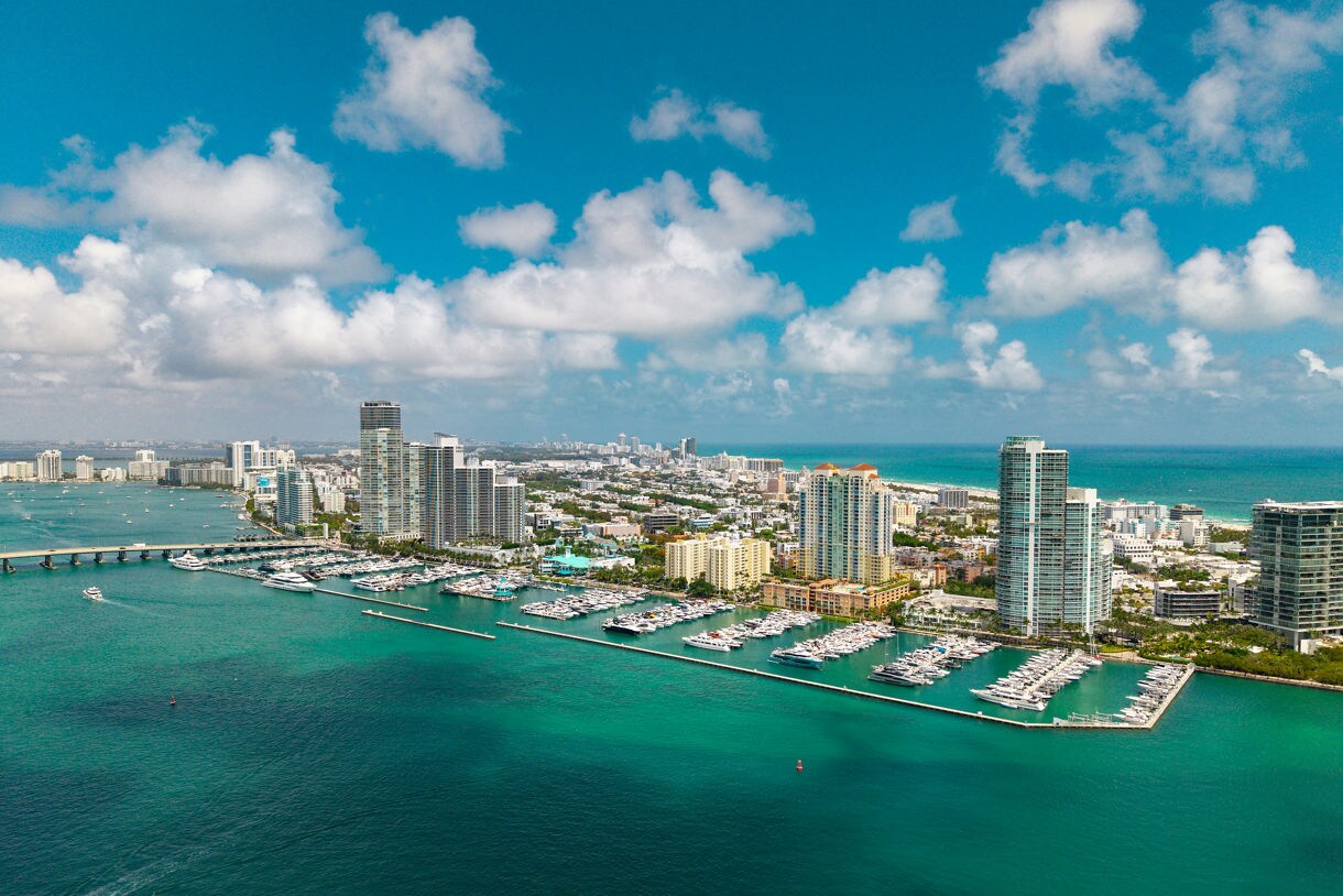 Aerial view of Miami’s skyline with high-rise buildings, marinas filled with yachts, and turquoise ocean waters under a partly cloudy sky.