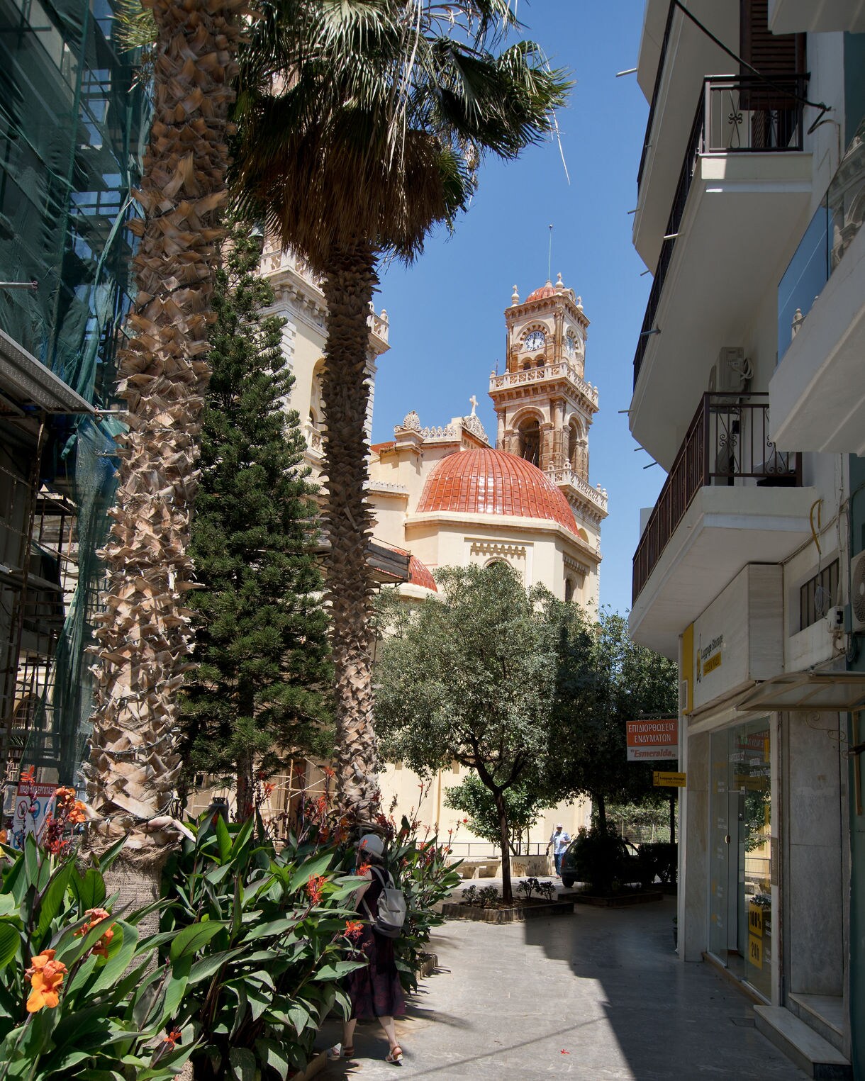Narrow pedestrian street in Heraklion lined with tall palm trees and flowering plants, leading toward the ornate Agios Minas Cathedral with its red dome and bell tower under a clear blue sky.