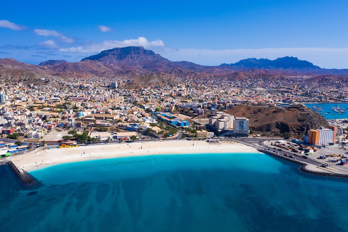 Aerial view of Mindelo, Cape Verde, featuring a bright turquoise bay, wide sandy beach, colorful city buildings and rocky mountains rising behind the town under a clear blue sky.