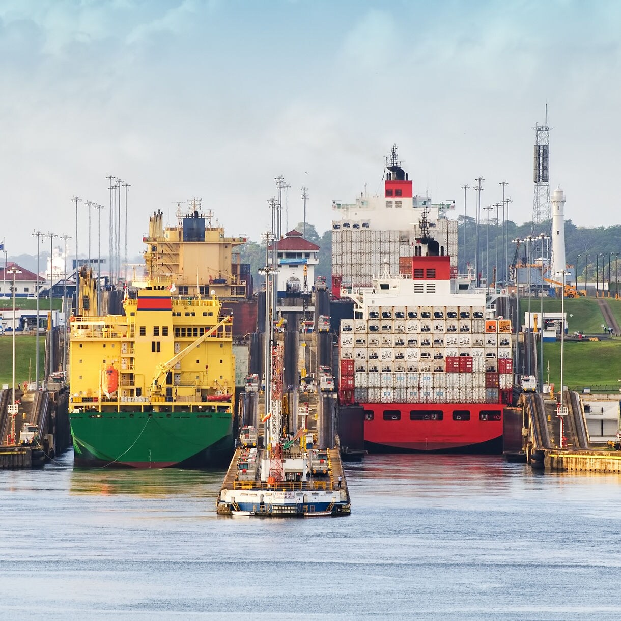 Two large cargo ships, one yellow and one red, passing side by side through the Miraflores Locks of the Panama Canal with surrounding buildings and greenery.