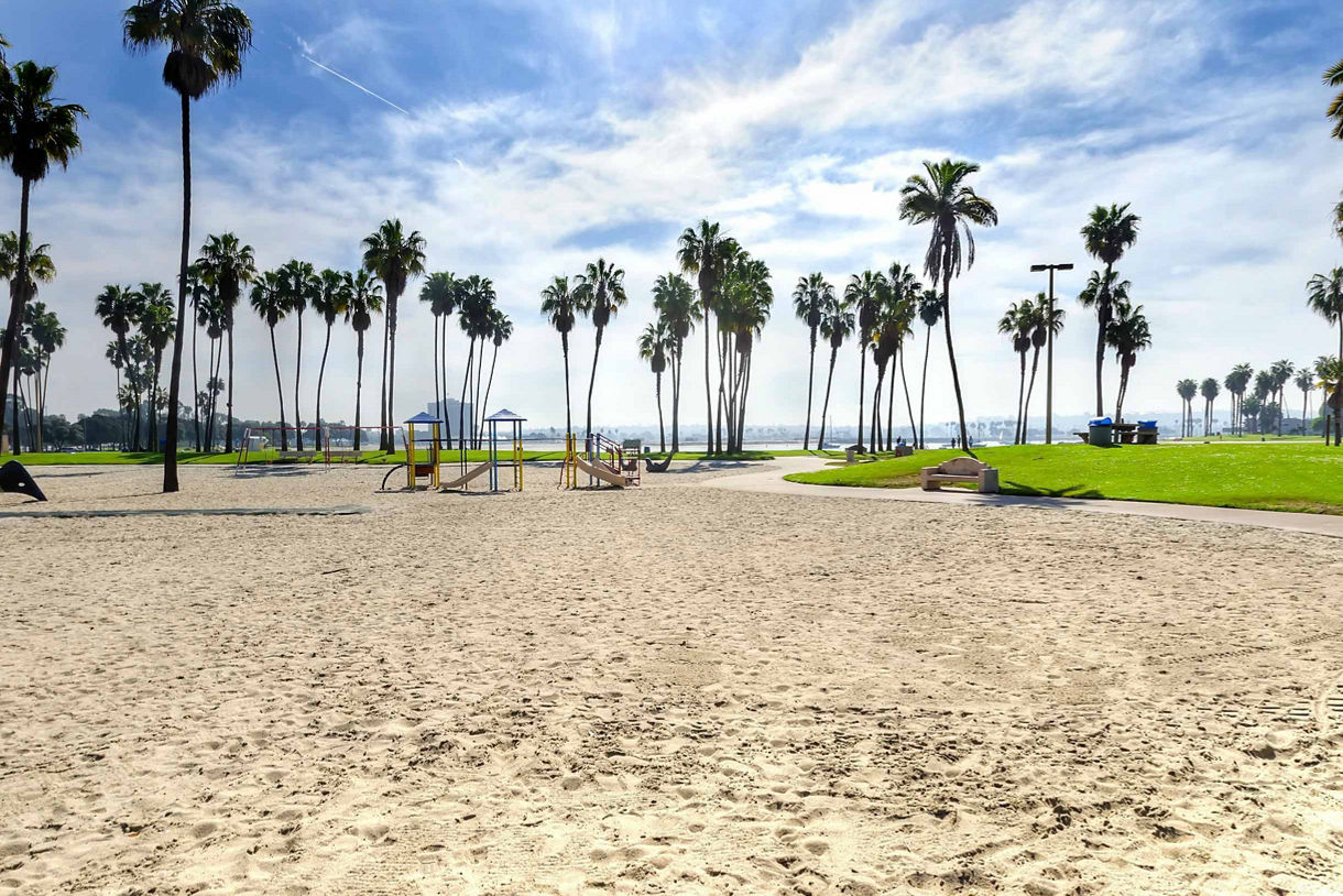 A sunny day at Mission Beach, featuring a sandy playground with yellow and blue play structures surrounded by tall palm trees.