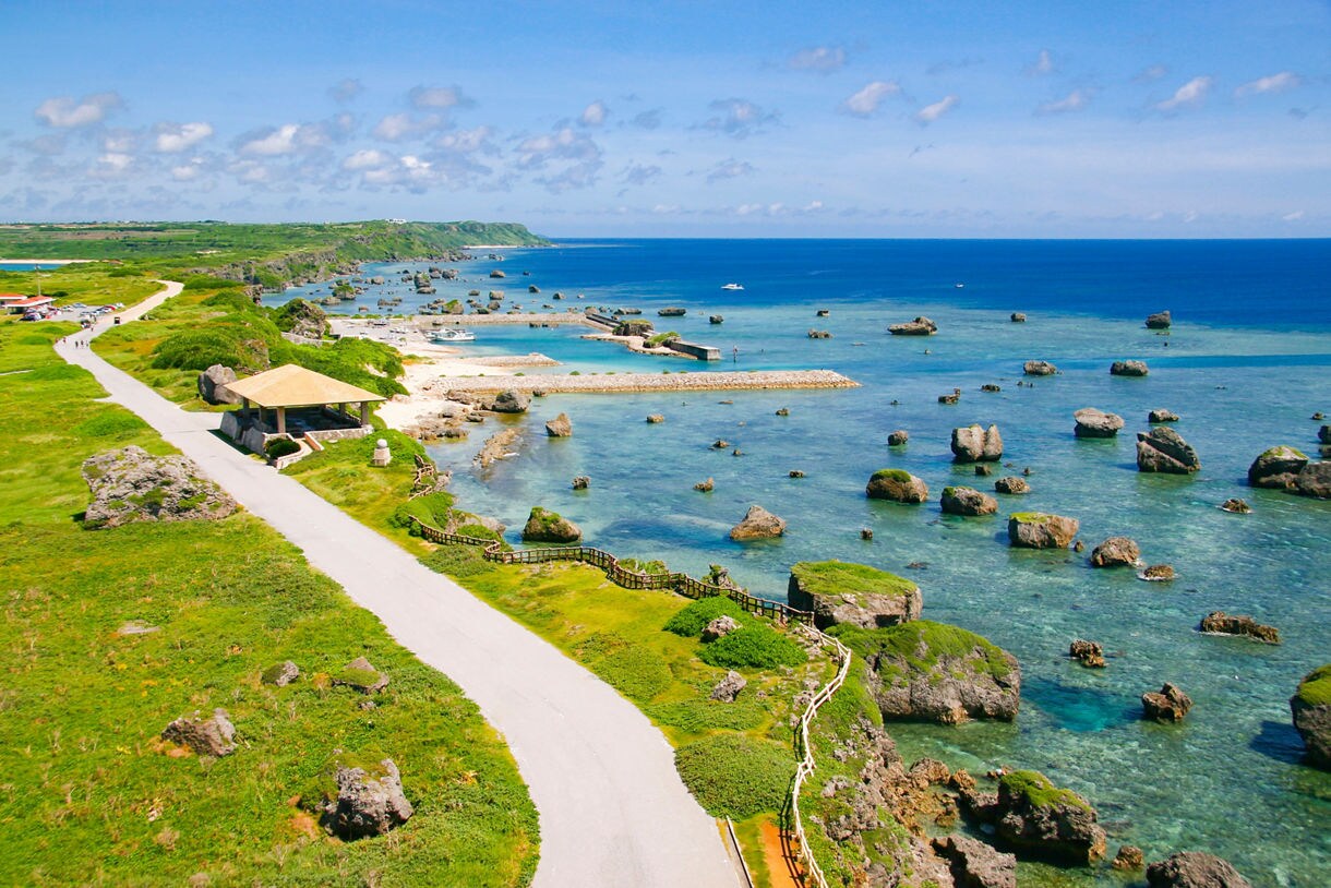 Elevated coastal view of Miyako Island featuring clear turquoise water dotted with large rock formations, a winding seaside road, green cliffs and a small pavilion overlooking the shoreline.