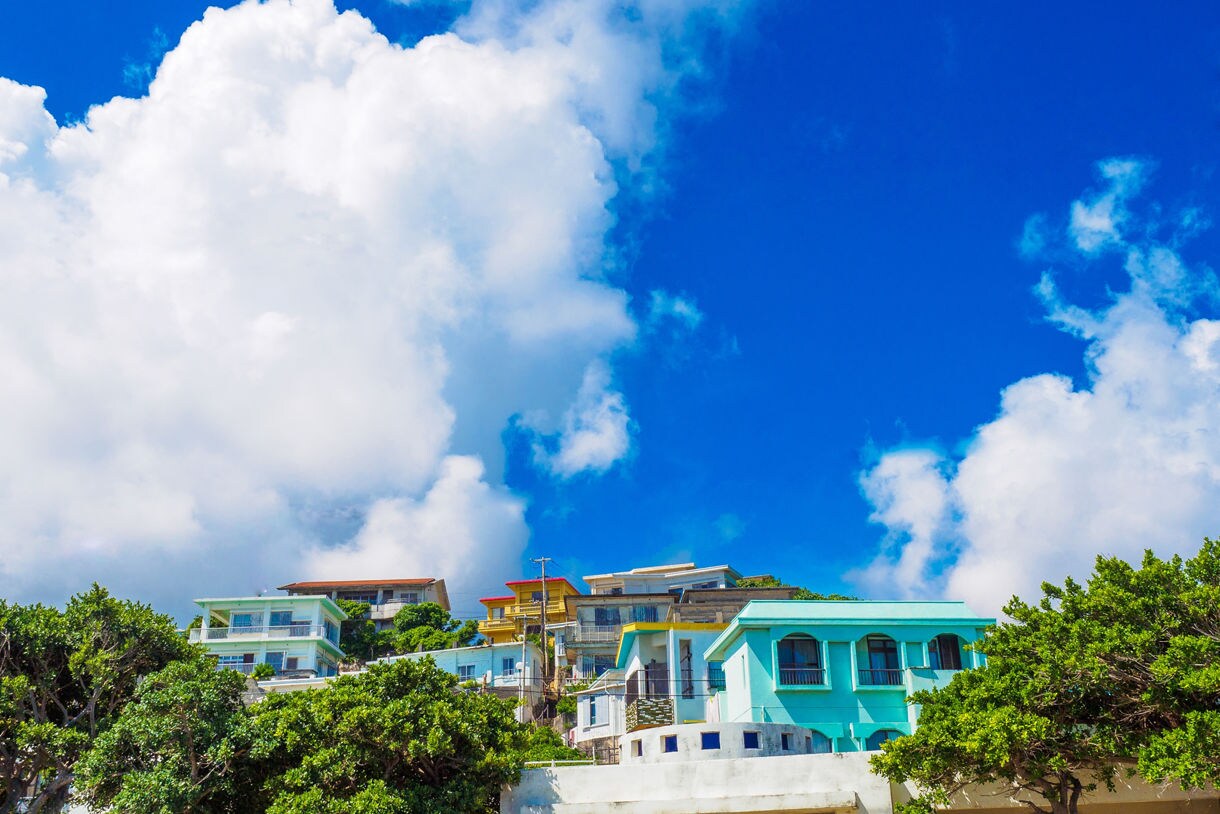 Row of bright, pastel-colored houses perched on a lush green hillside in Miyakojima, set against a vibrant blue sky filled with large white clouds.