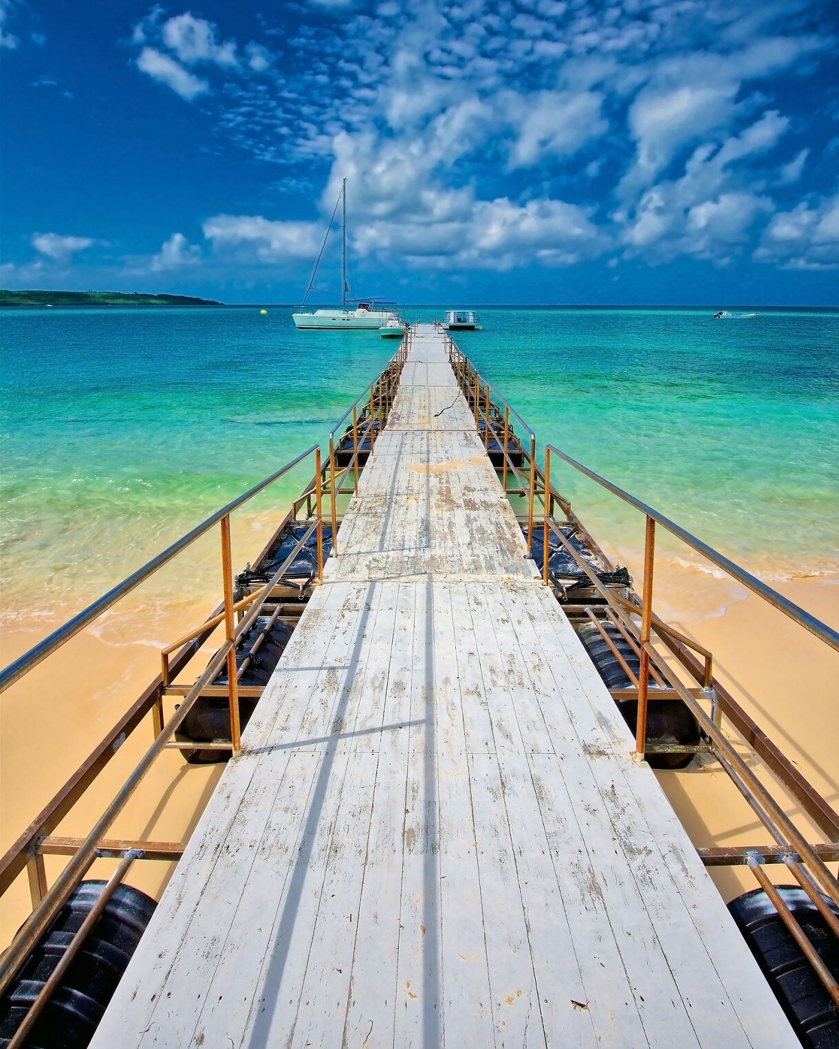 Long wooden pier extending into bright turquoise water on Miyakojima Island, with a sailboat anchored offshore under a vivid blue sky filled with scattered clouds.