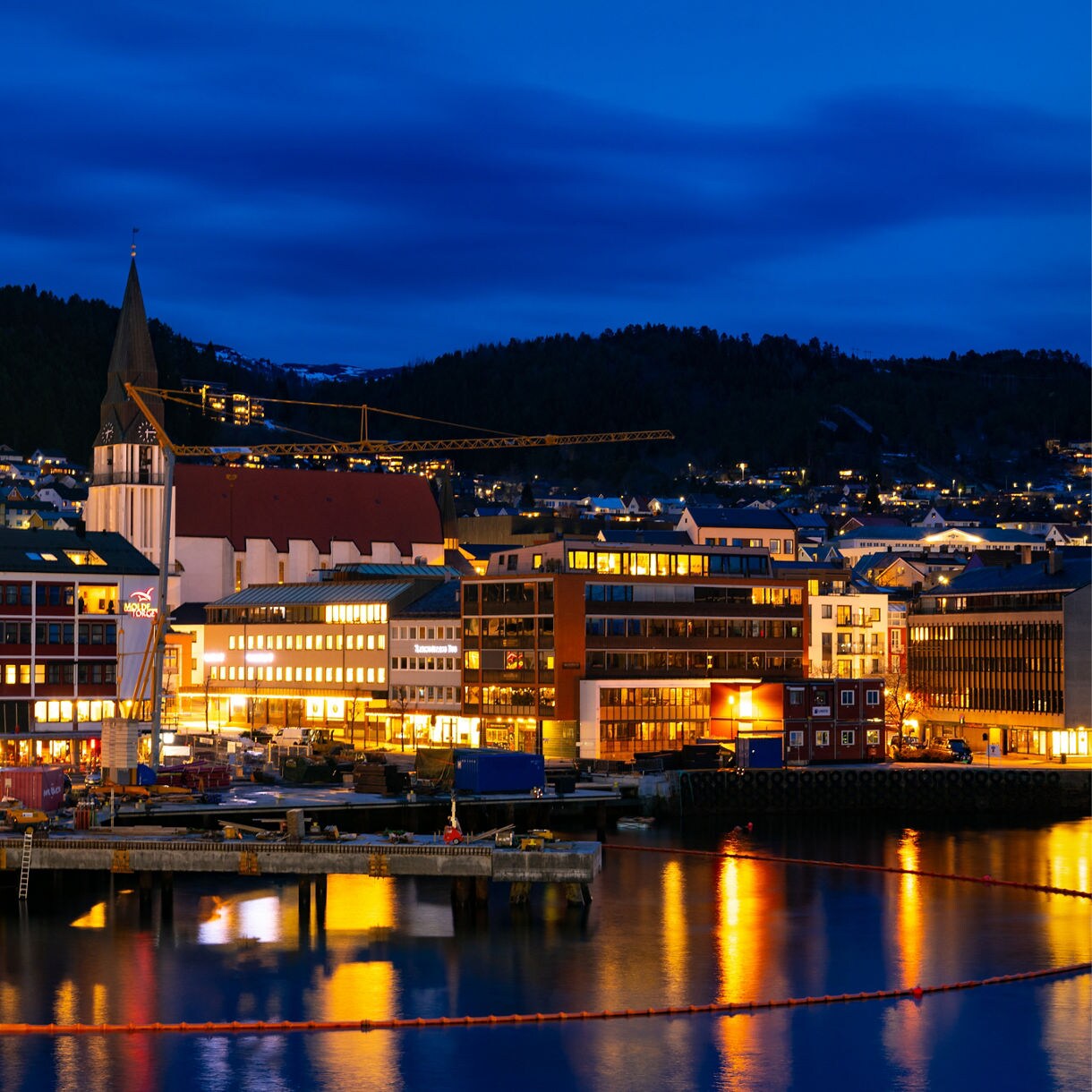 Nighttime view of Molde, Norway, with illuminated buildings reflecting on the harbor water, a church spire rising in the center and hills dotted with lights under a deep blue sky.