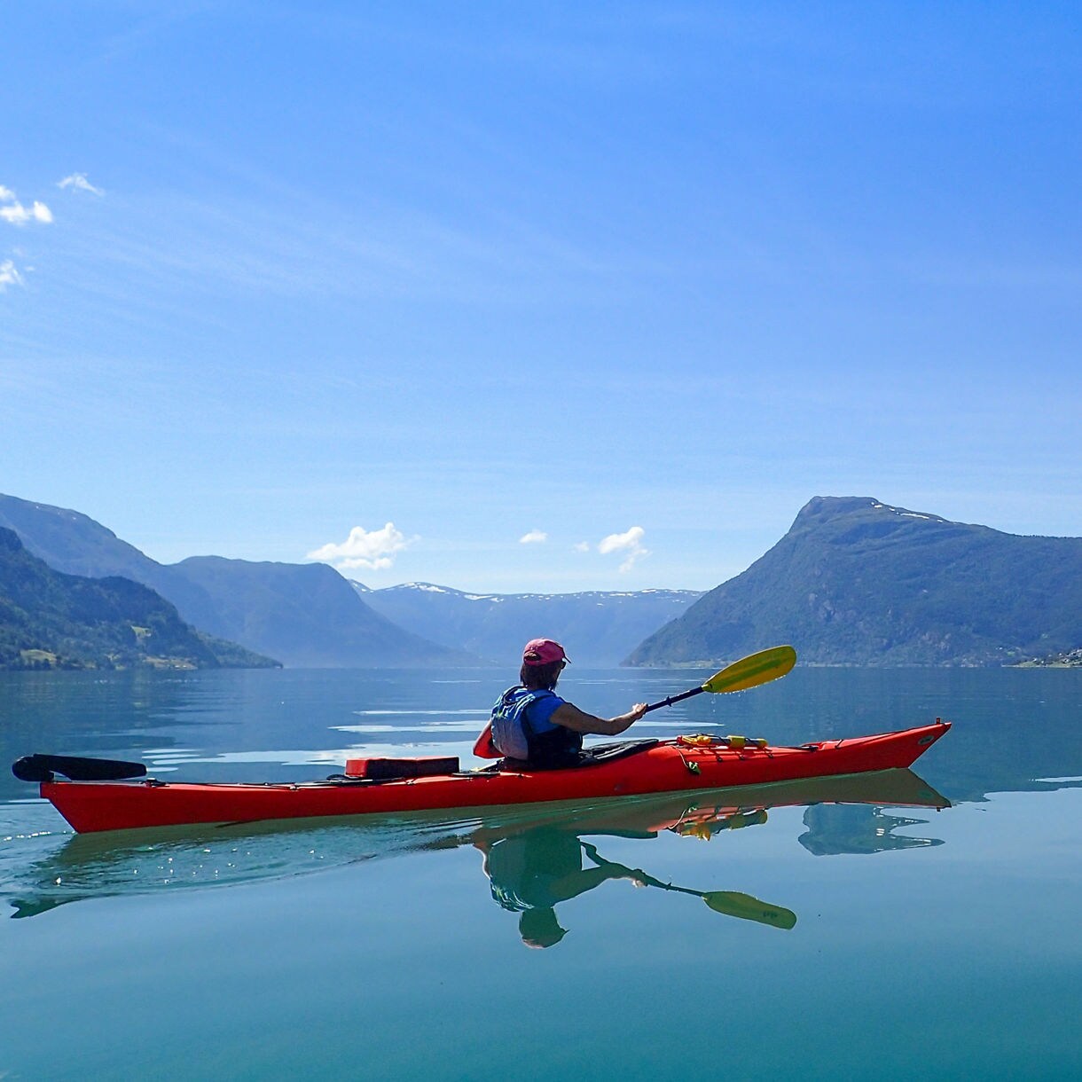 A kayaker in a red kayak paddling on calm, reflective fjord water surrounded by tall mountains under a bright blue sky in Norway.