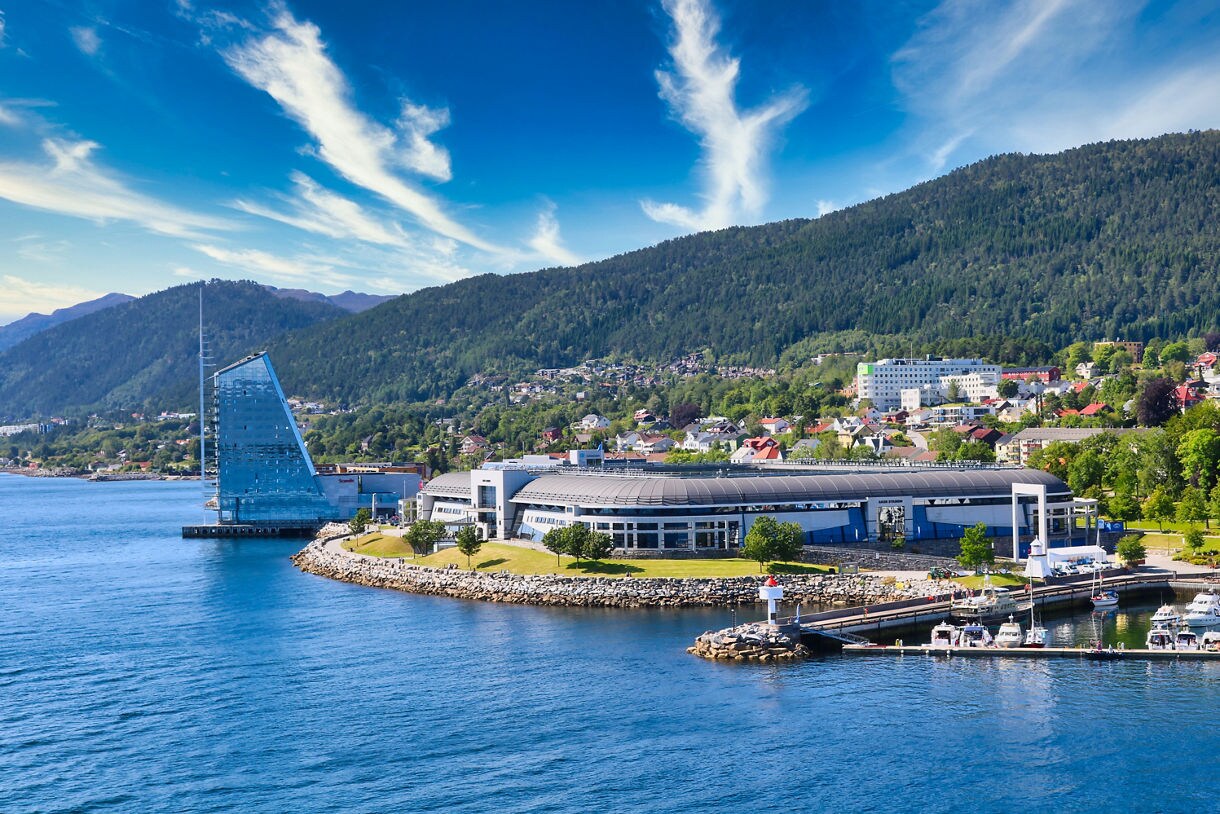 A waterfront scene in Molde, Norway, showing a modern glass building shaped like a sail, a curved convention center, small boats at a marina and forested hills rising behind the town under a bright blue sky.