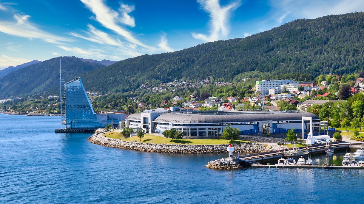 A waterfront scene in Molde, Norway, showing a modern glass building shaped like a sail, a curved convention center, small boats at a marina and forested hills rising behind the town under a bright blue sky.