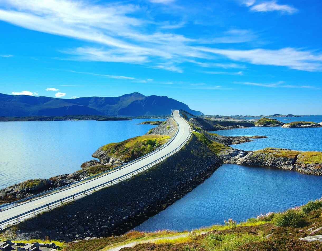 A striking stretch of the Atlantic Ocean Road in Norway, curving over low rocky islands surrounded by bright blue water, with distant mountains under a clear sky.