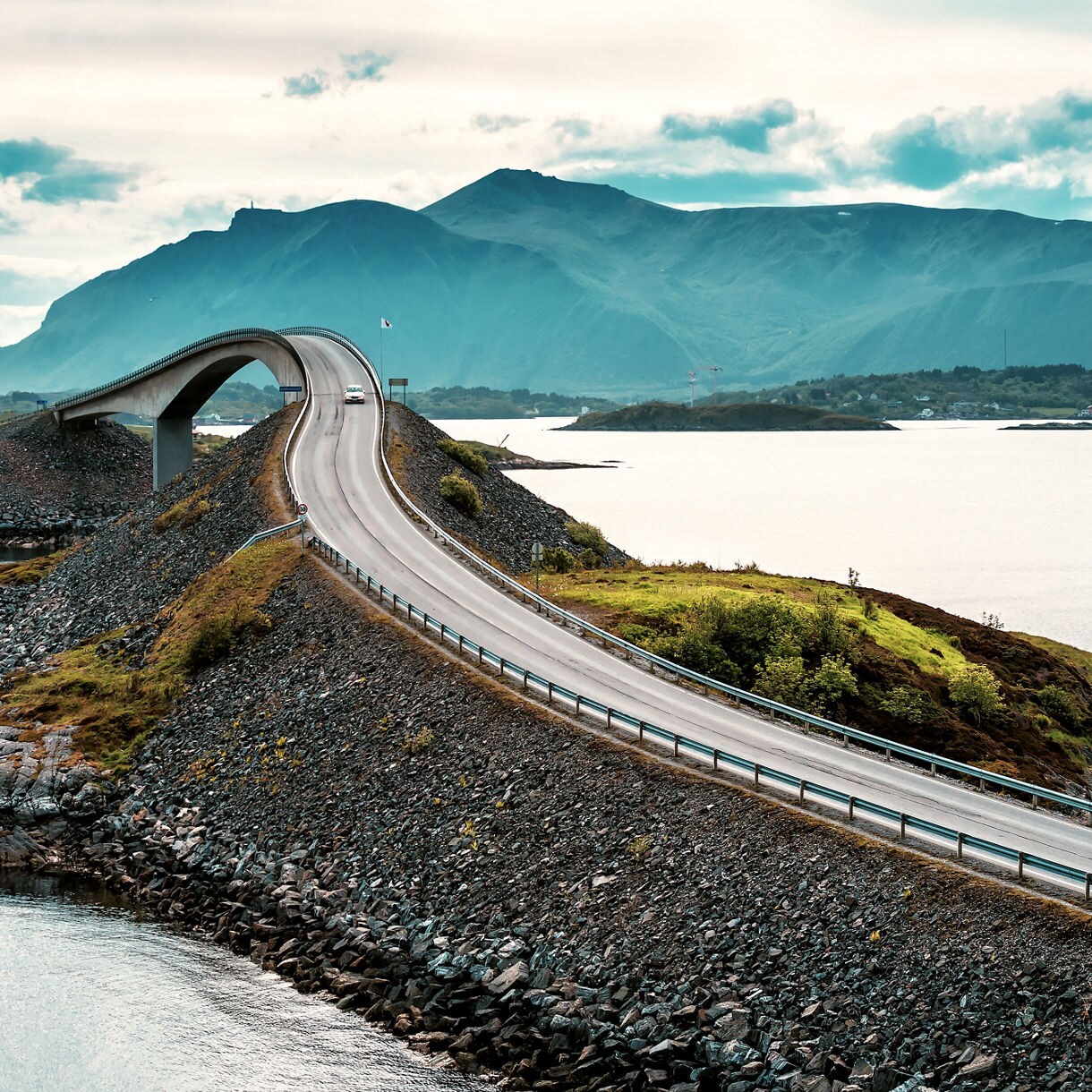 A dramatic stretch of Norway’s Atlantic Ocean Road curving over a raised causeway above rocky shores, with distant mountains and calm water under a cloudy sky.