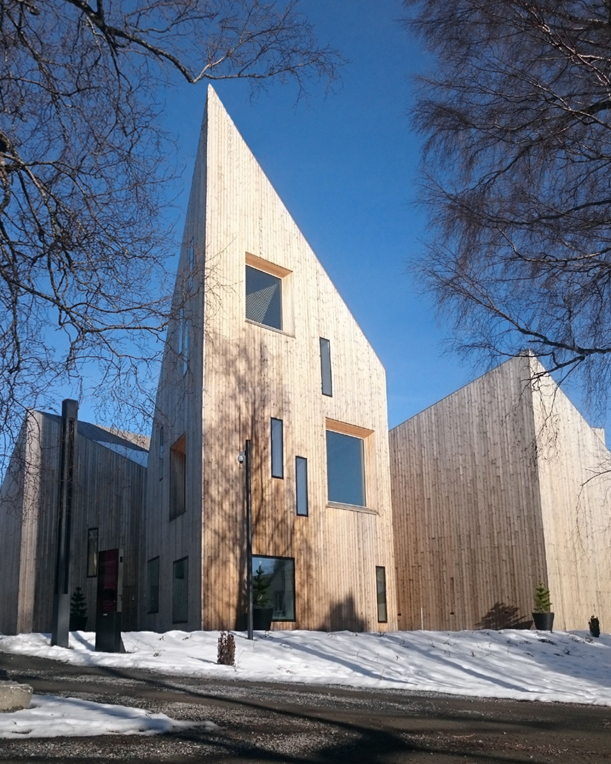 A contemporary wooden building with tall angular rooflines and narrow windows at the Romsdal Museum in Molde, Norway, surrounded by snow and bare winter trees under a bright blue sky.