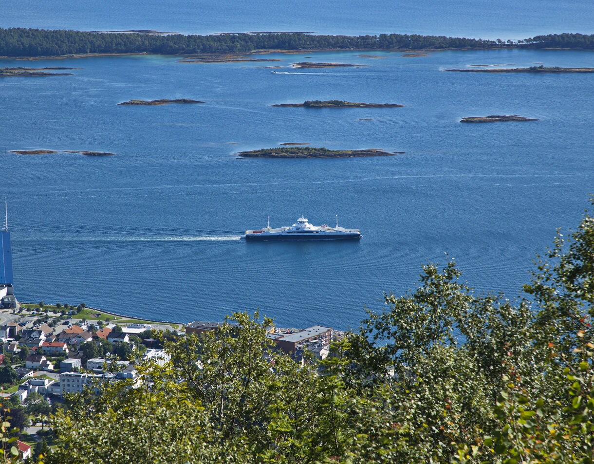 High vantage view overlooking Molde, Norway, with leafy trees in the foreground, a ferry crossing bright blue fjord waters and scattered low islands under a clear sky.