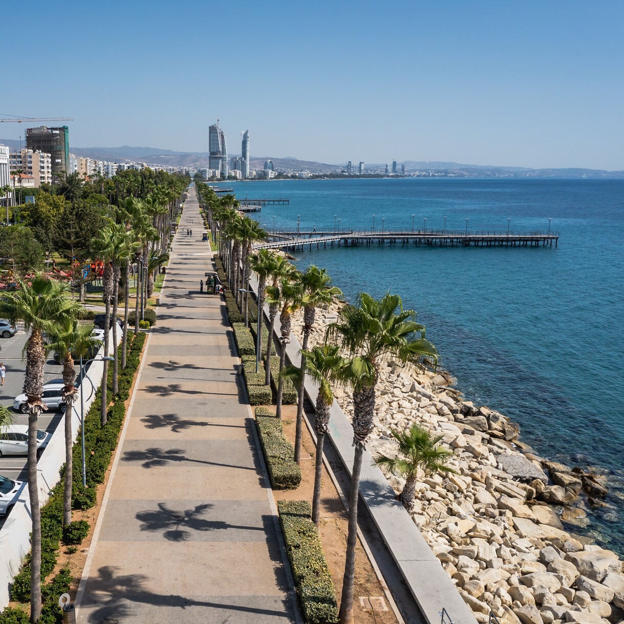 Aerial view of Limassol’s Molos Promenade lined with palm trees beside clear blue water, with parks, walkways and modern city buildings stretching into the distance.