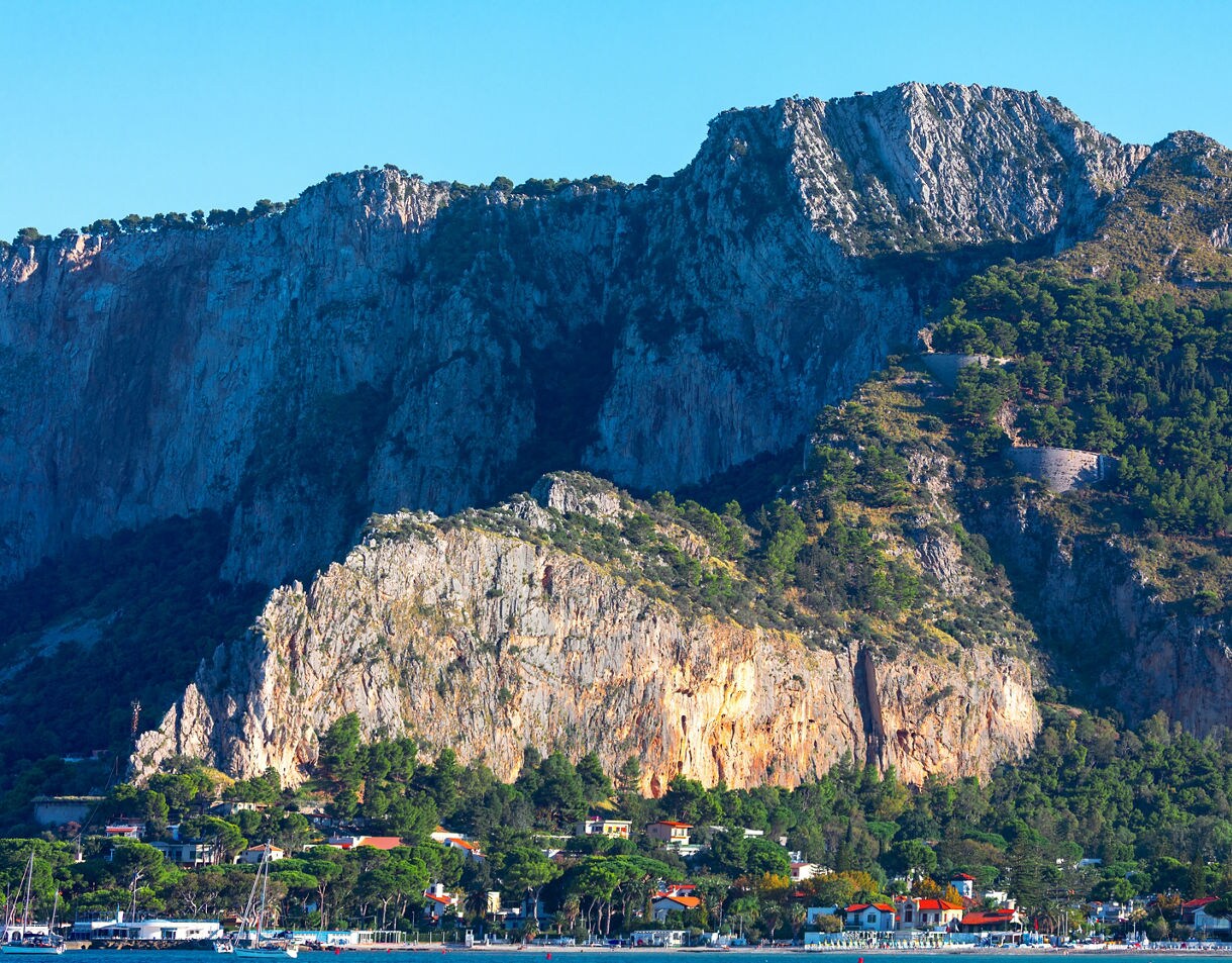 A coastal village at the base of steep rocky cliffs in Mondello, Sicily, with clusters of homes surrounded by pine trees near the blue shoreline.