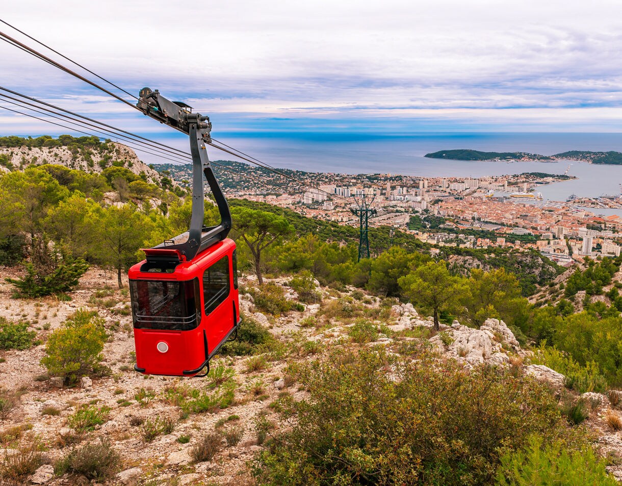 Red Mont Faron cable car suspended over rocky hills and green pines with a panoramic view of Toulon, its harbor and the Mediterranean Sea in the distance.