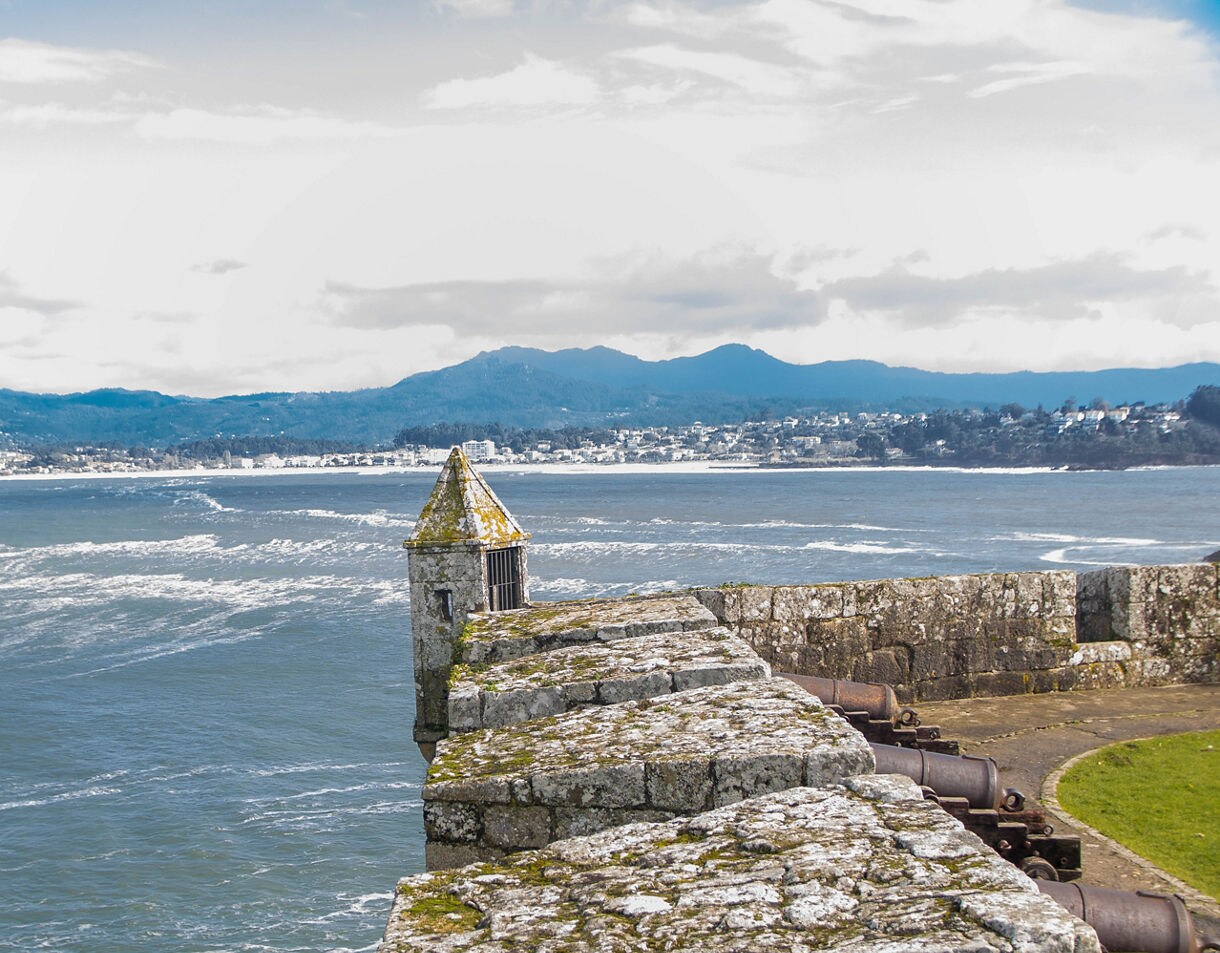 Stone ramparts of Monte Real Fortress overlooking a choppy blue bay, with an old watchtower and cannons along the wall and mountains visible across the water.