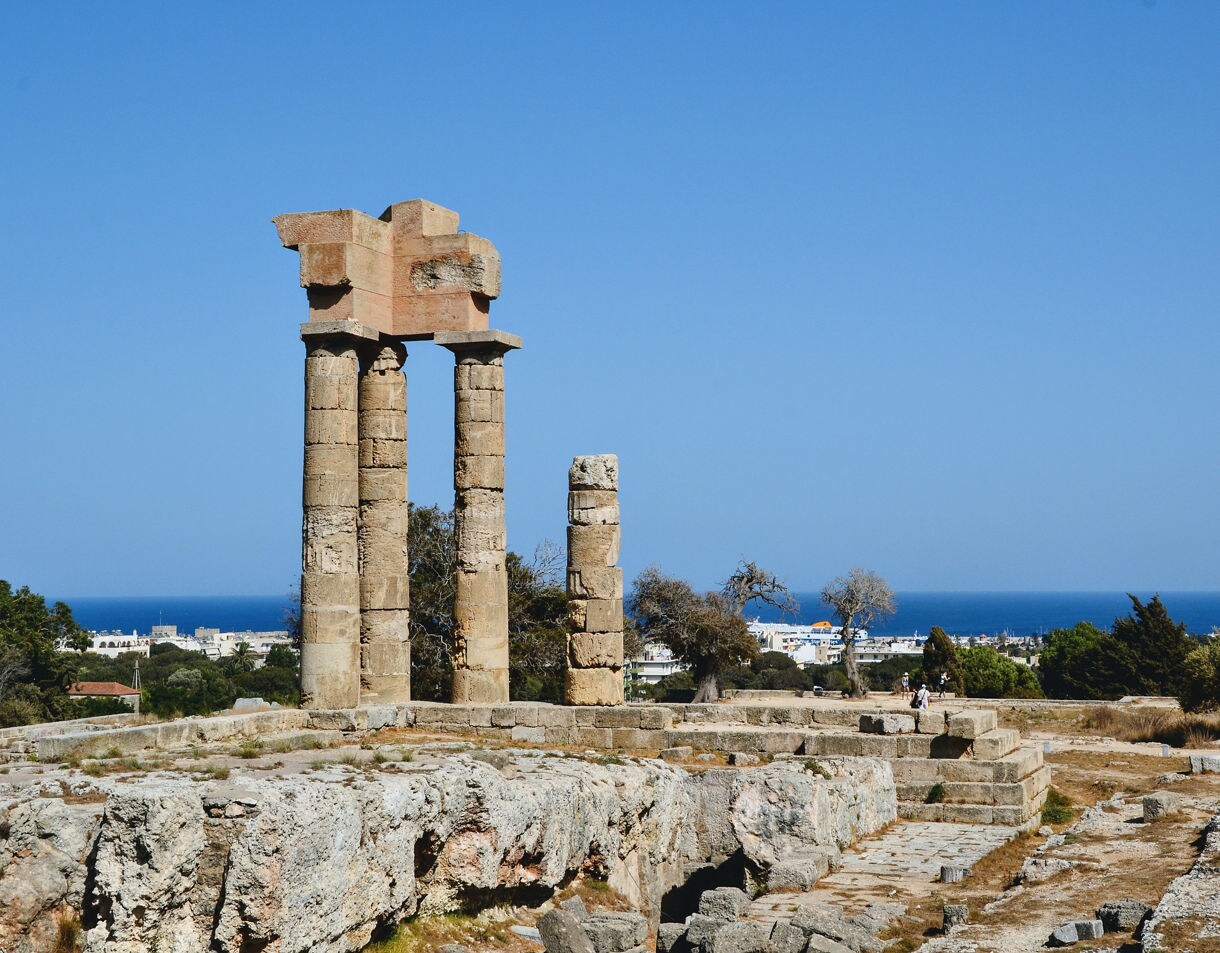 Weathered stone columns of the ancient Acropolis of Rhodes on Monte Smith Hill overlooking the city and sea under a clear blue sky.