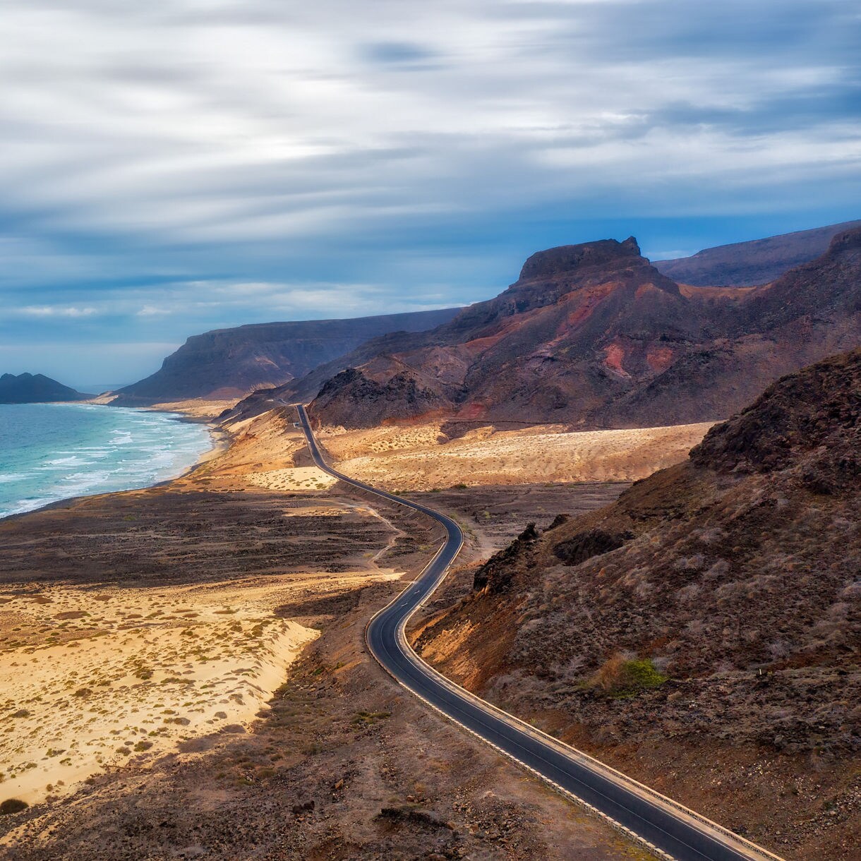 Panoramic view of a winding coastal road cutting through dry volcanic terrain beside a long stretch of turquoise ocean, with rugged cliffs and layered mountains rising under a cloudy sky.