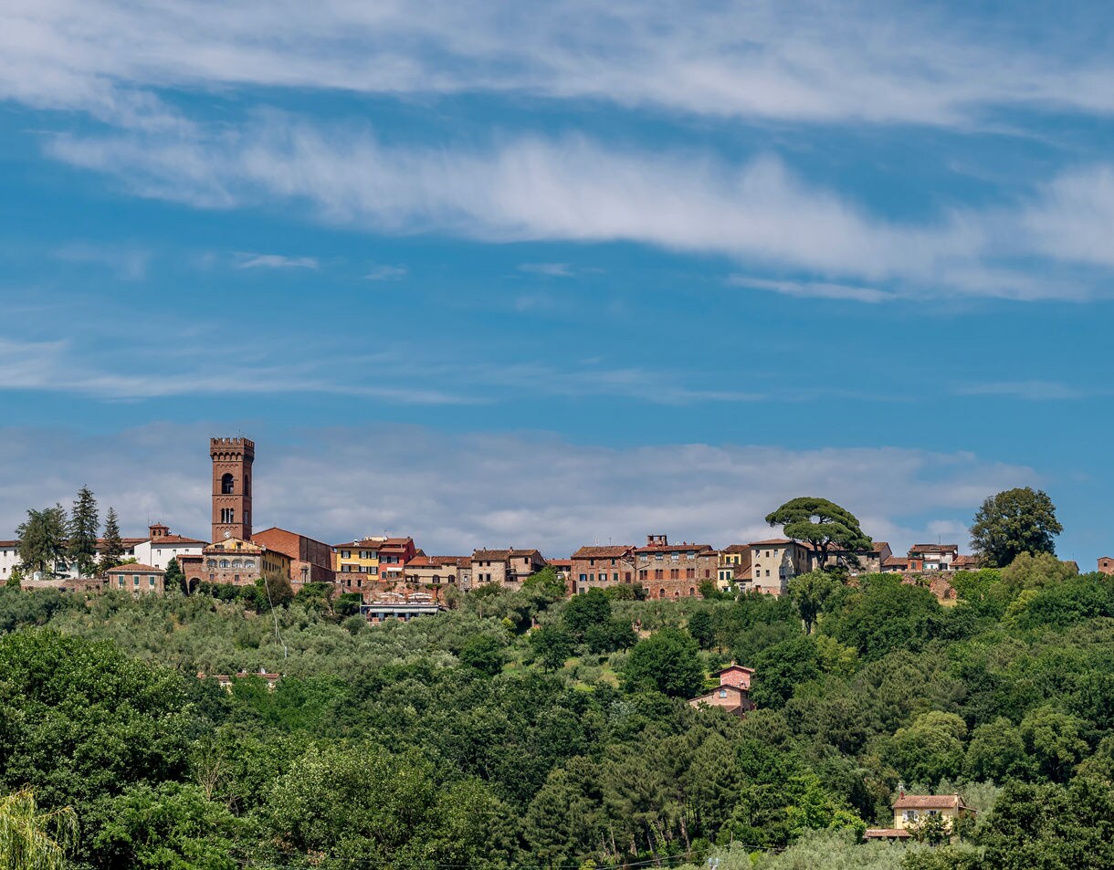 A hilltop village in Tuscany with stone buildings and a tall brick tower, surrounded by green trees and olive groves under a blue sky with wispy clouds.