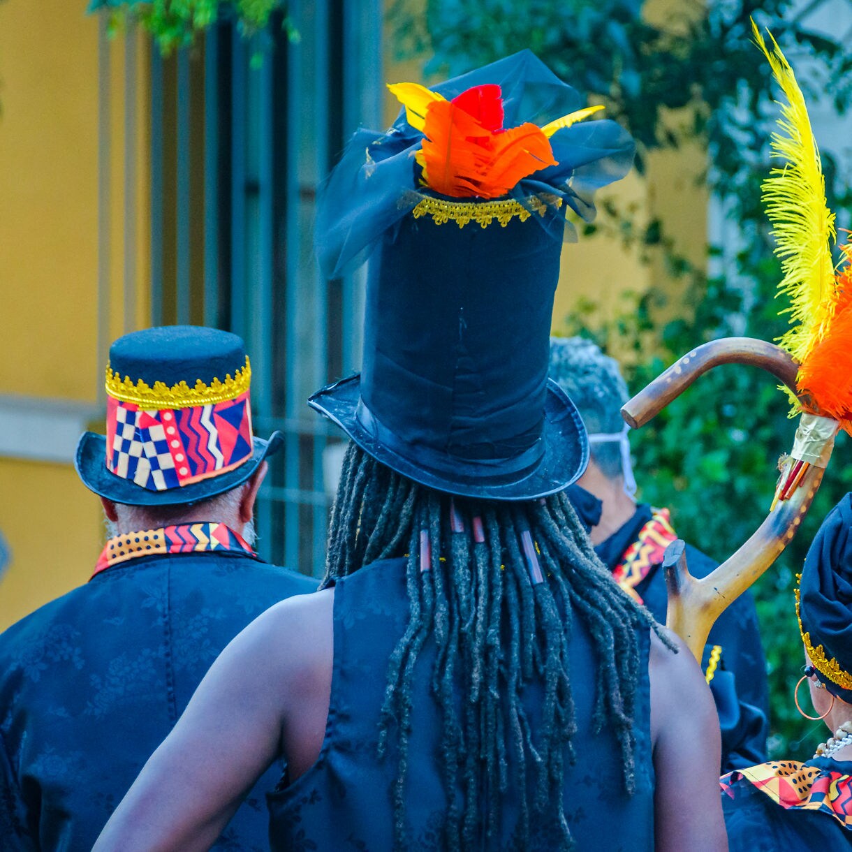 Group of performers seen from behind wearing black outfits with colorful patterned details, tall hats, headwraps and bright feather plumes during a carnival celebration.