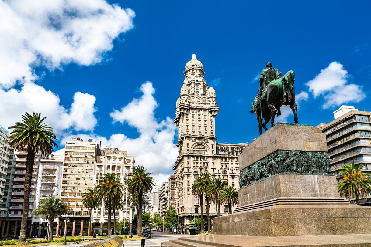 A lively city plaza in Montevideo featuring an ornate historic tower, modern buildings, tall palm trees and a large equestrian statue on a stone pedestal under a bright blue sky.