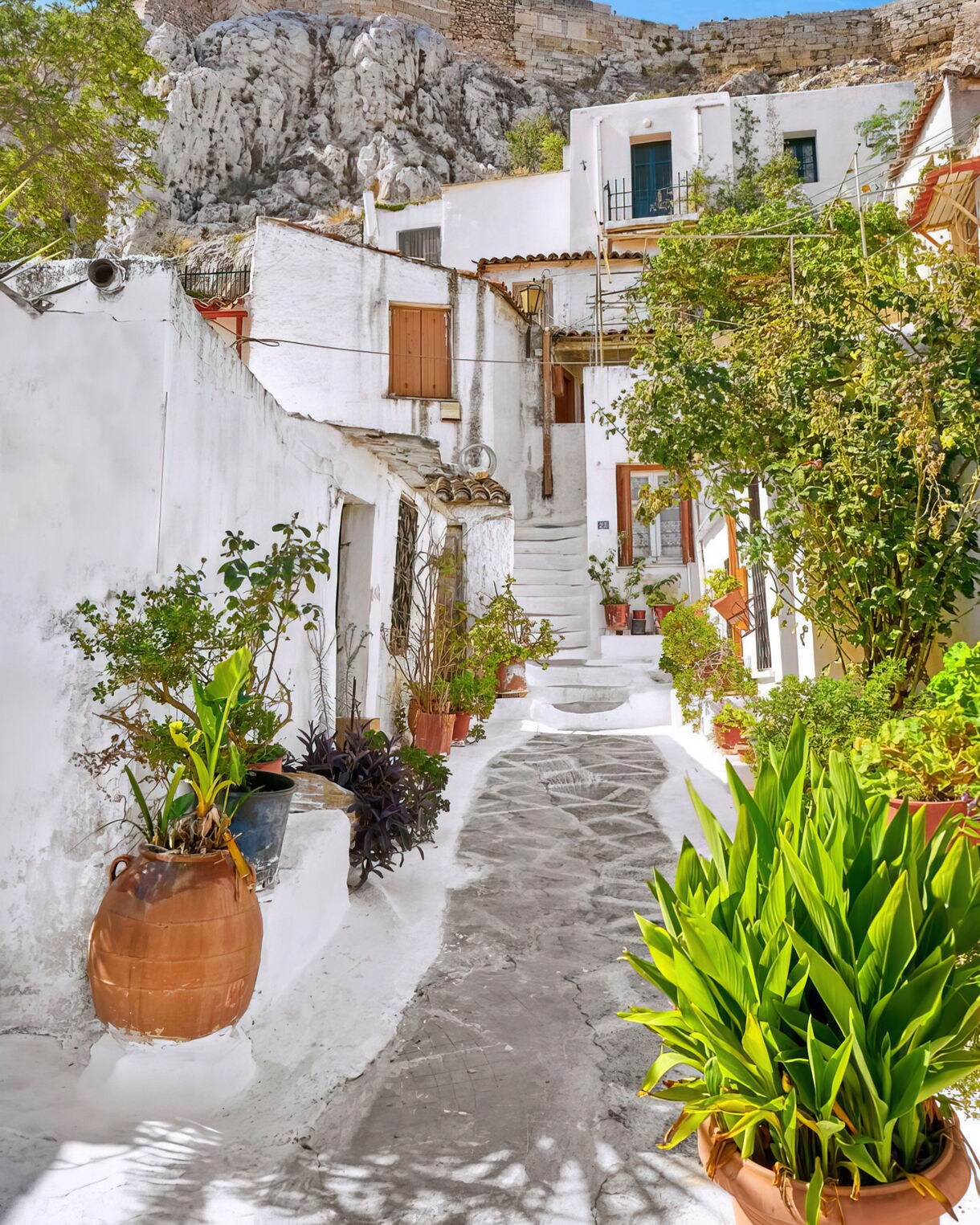 Narrow walkway lined with white buildings and potted plants leading up toward rocky historic ruins.
