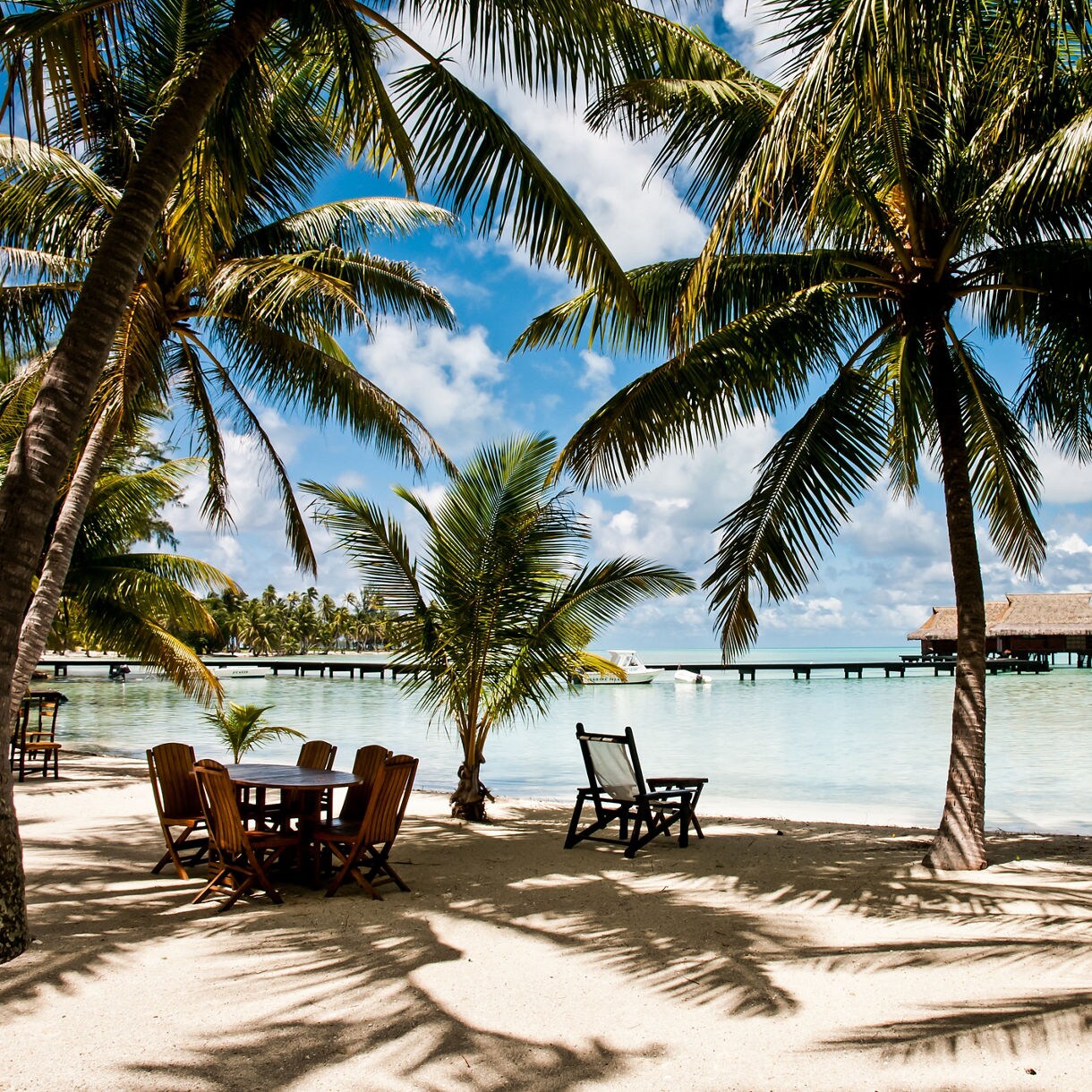 Sandy beach shaded by palm trees with wooden chairs and tables, overlooking a calm lagoon and an overwater bungalow in the distance.