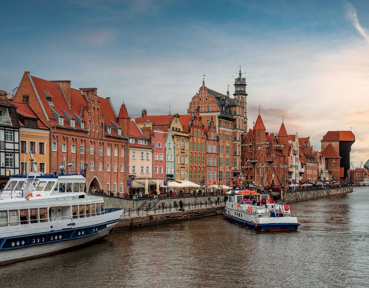 View of the Motława River in Gdańsk, Poland, with colorful historic buildings lining the waterfront, boats docked along the embankment and the medieval Crane Gate visible in the distance at sunset.