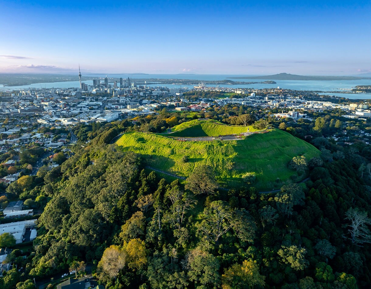 Aerial view of Mount Eden’s grassy crater with Auckland city skyline and harbor in the background.