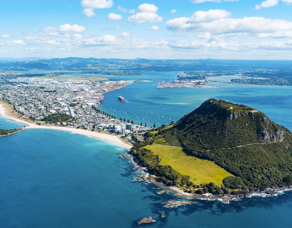 Aerial view of Mount Maunganui with a grassy volcanic hill, white sandy beaches, turquoise ocean and the city of Tauranga stretching inland.