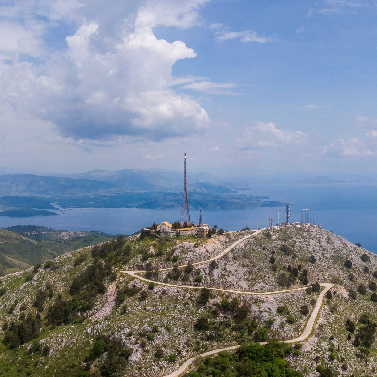 Aerial view of Mount Pantokrator in Corfu with winding roads, rocky slopes, antennas on the summit and the blue sea stretching into the horizon.