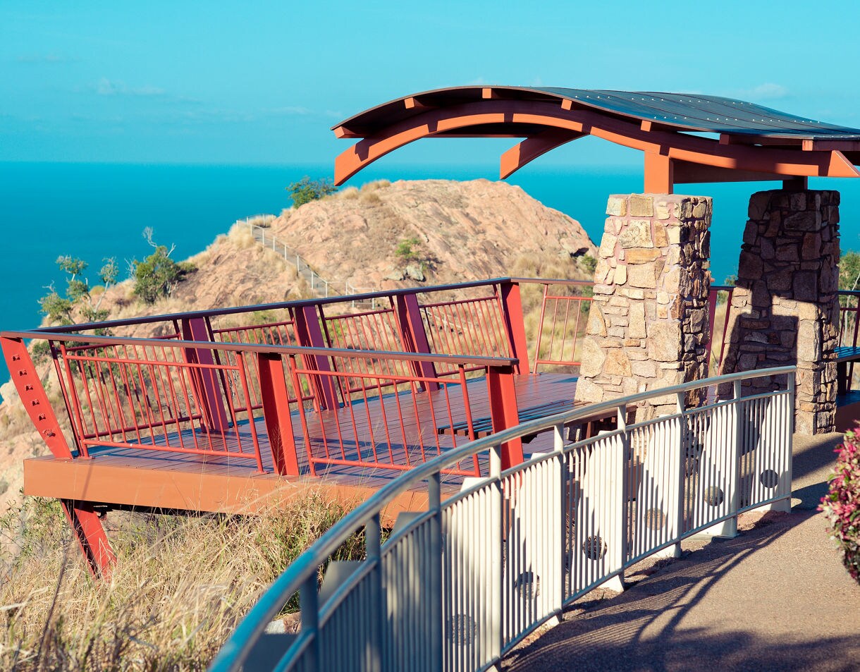 A red metal and stone lookout platform on a hilltop overlooking bright blue ocean under clear skies.