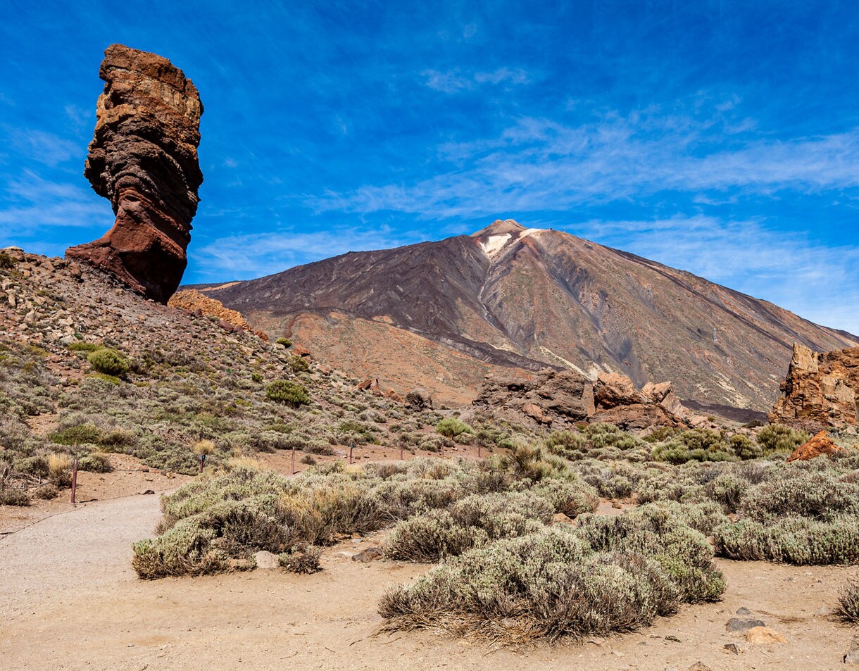 View of Mount Teide in Tenerife, Spain, with rugged volcanic rock formations and desert shrubs under a vivid blue sky.