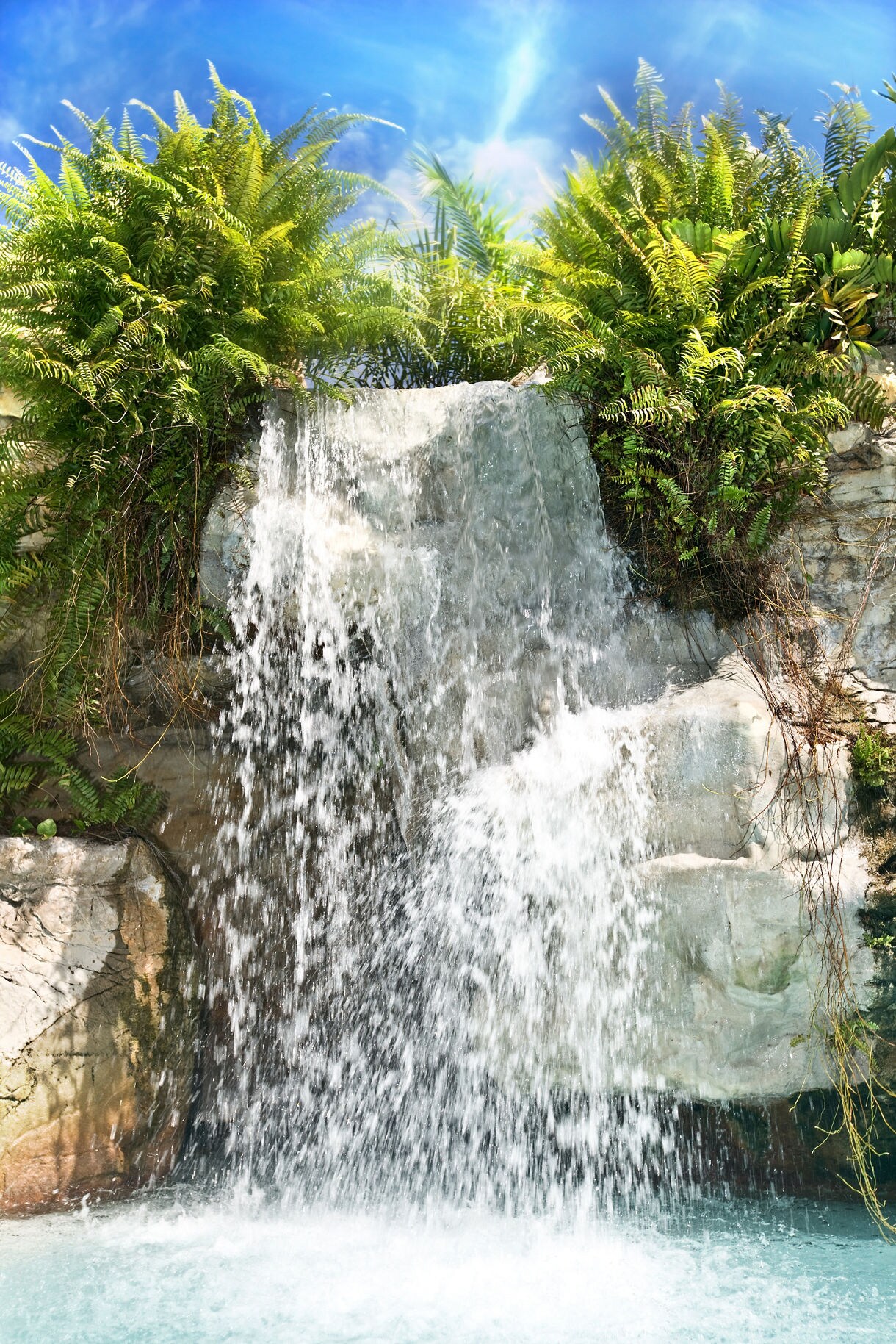 Tropical waterfall spilling over rocky ledges surrounded by ferns and palms under a bright blue sky.