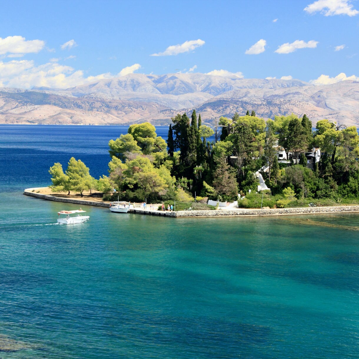 Small tree-covered Mouse Island near Corfu, surrounded by turquoise and deep blue sea with distant mountains in the background.