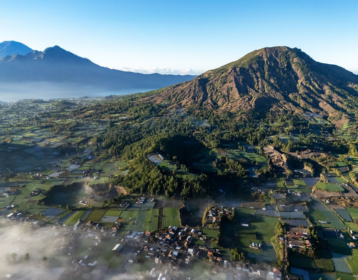 Aerial view of Mount Batur and the surrounding valley, featuring terraced farmland, scattered homes, drifting morning mist and distant mountain ridges under clear blue sky.