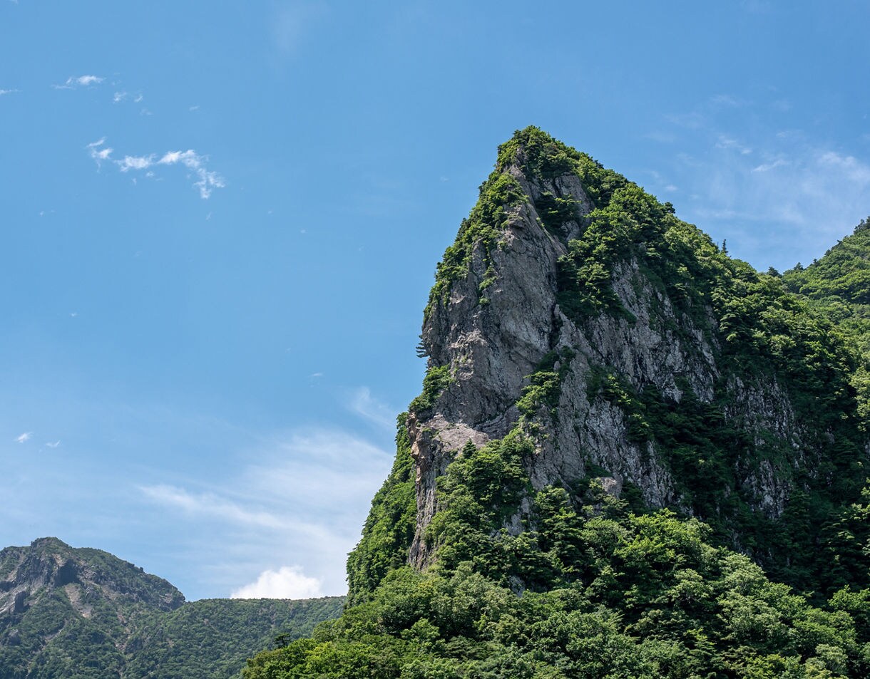 Jagged rocky peak of Mount Halla on Jeju Island, South Korea, rising above dense green forest under a bright blue sky.