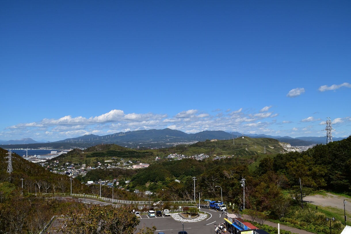 Wide panoramic overlook showing rolling green hills, clusters of houses, distant mountains and wind turbines, with a curved parking area and vehicles in the foreground under a clear sky with scattered clouds.