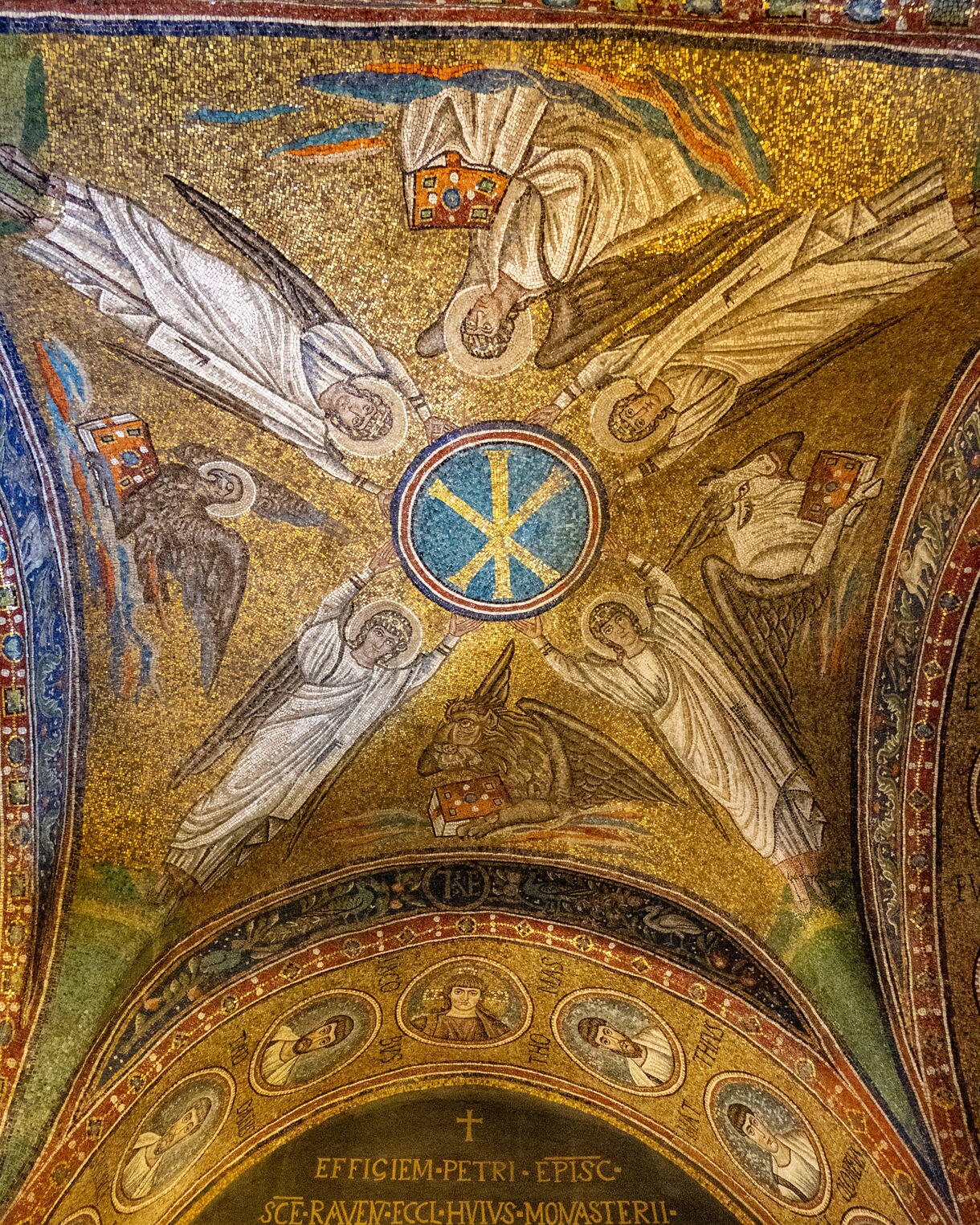 Golden Byzantine mosaic ceiling inside the Archiepiscopal Chapel in Ravenna featuring angels surrounding the Chi-Rho symbol above an arched inscription.