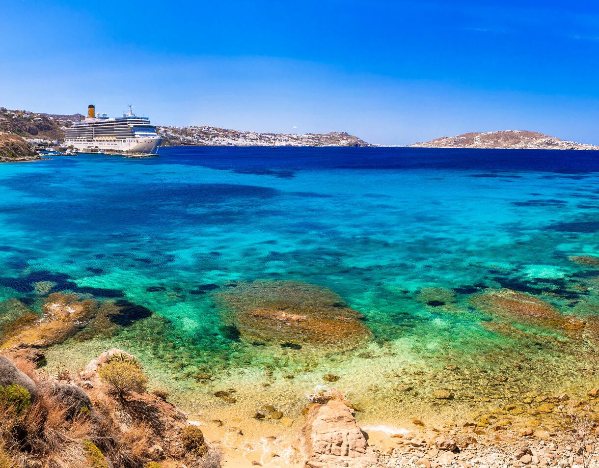 Panoramic view of a Mykonos beach with turquoise sea, sandy shoreline, hillside whitewashed houses and a cruise ship anchored offshore.