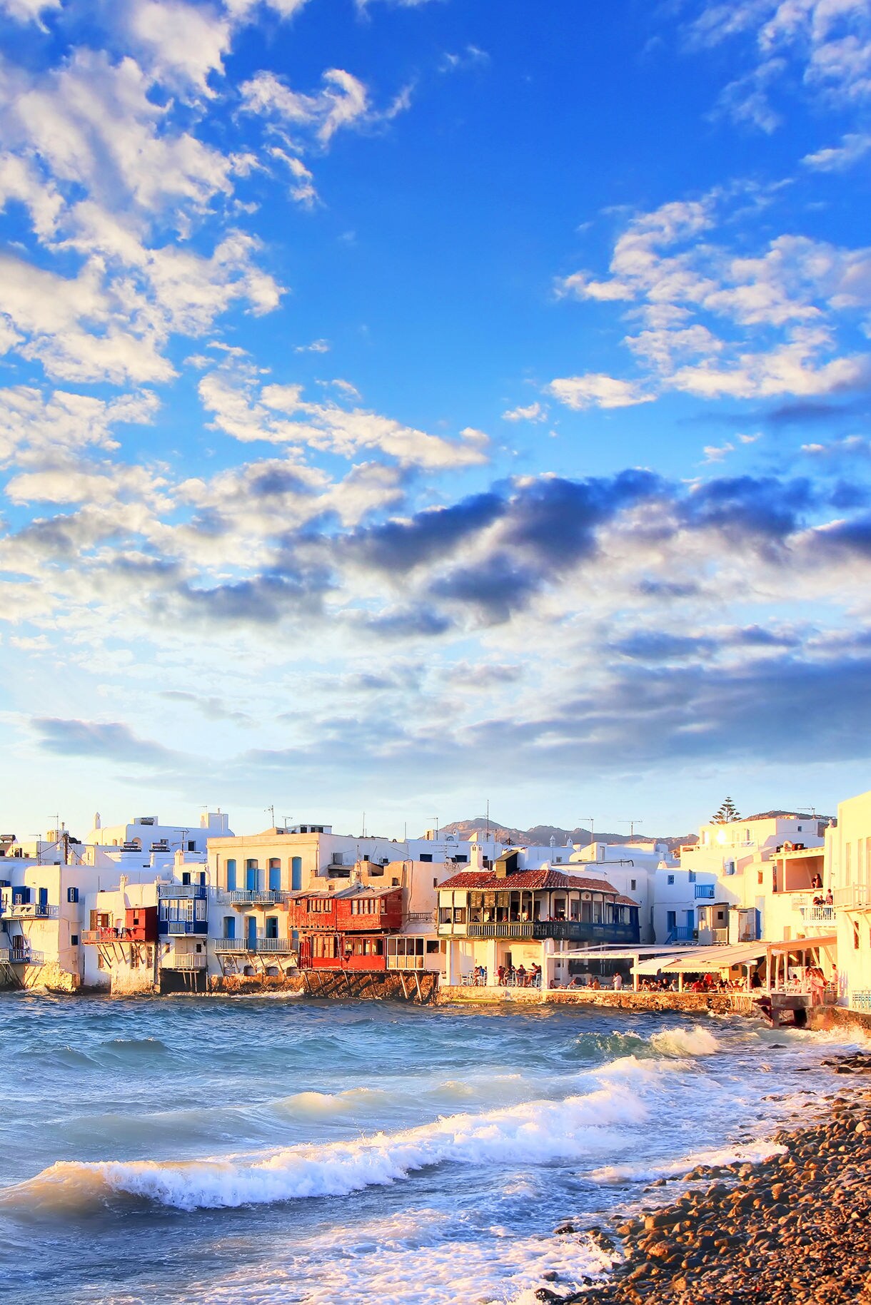 Sunset view of Little Venice in Mykonos with whitewashed buildings and colorful balconies along the waterfront as waves roll onto the shore.