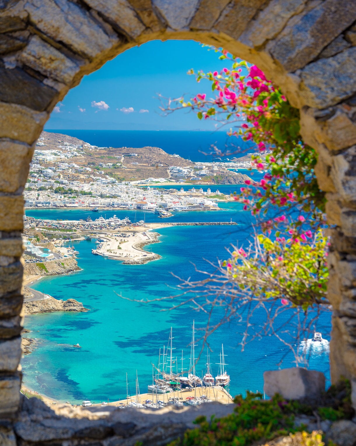 View of Mykonos coastline with turquoise waters, sailboats and white buildings, framed by a stone arch with pink flowers.