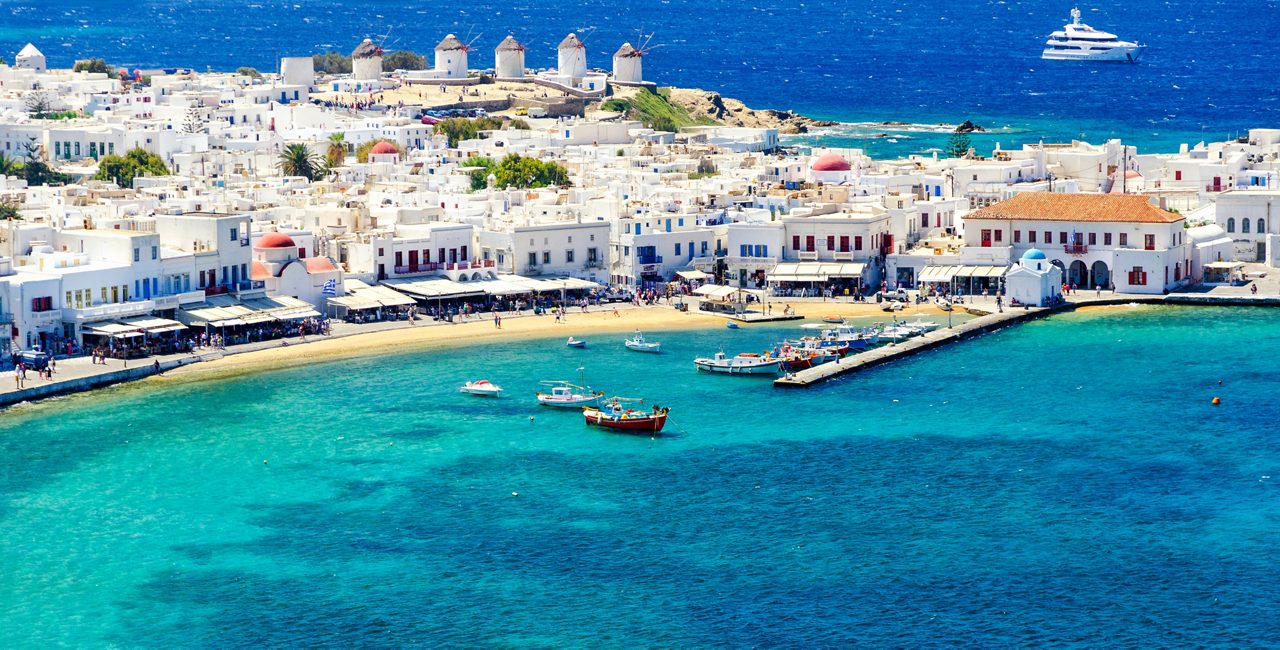 Aerial view of Mykonos waterfront with whitewashed buildings, red-domed chapels, traditional windmills, and boats anchored in clear turquoise water.