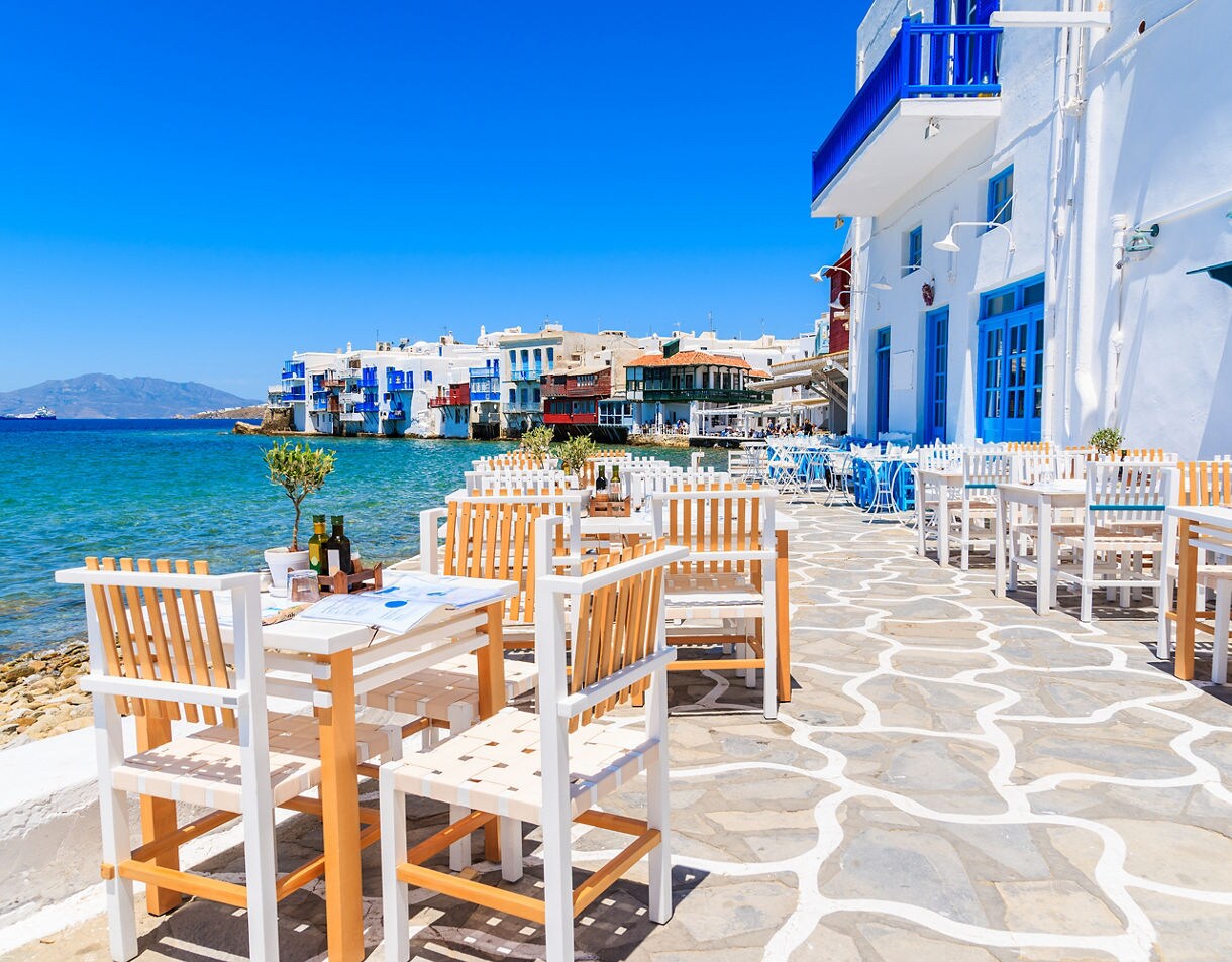 Outdoor waterfront restaurant with white wooden tables and chairs in Mykonos’ Little Venice, overlooking colorful seaside houses with blue balconies above the Aegean Sea.