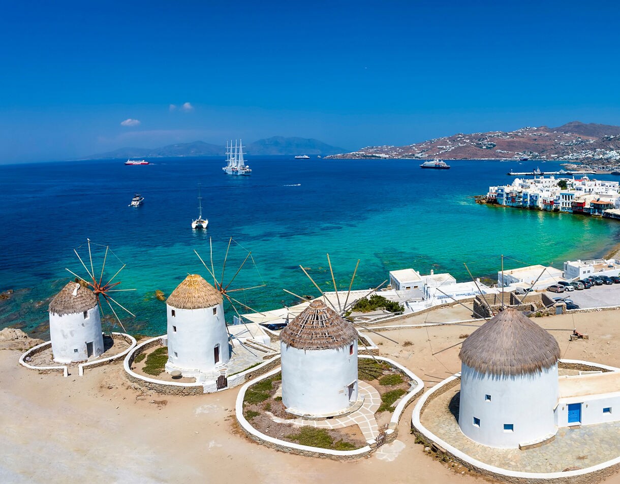 Aerial view of Mykonos Town with traditional white windmills overlooking turquoise waters, boats and whitewashed houses along the coast.