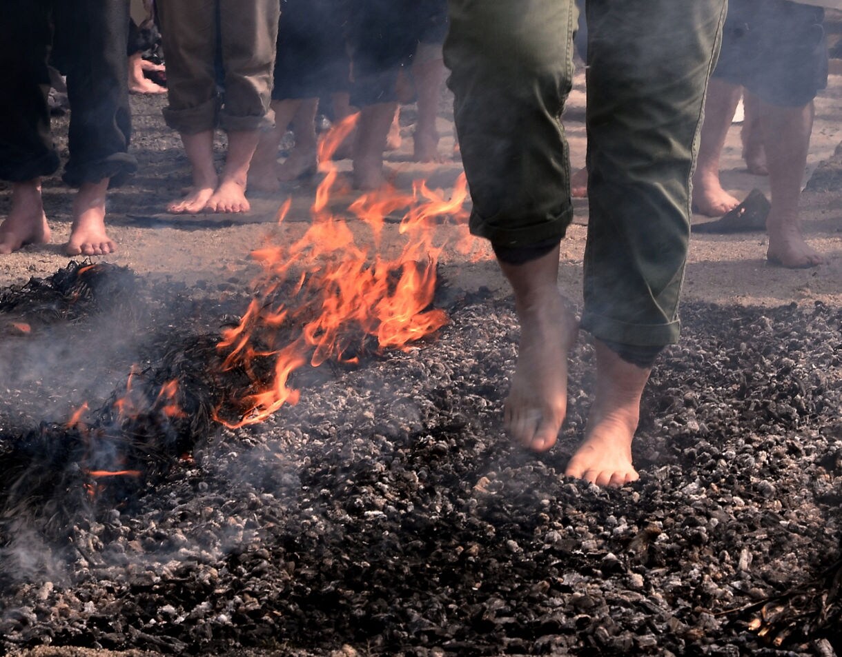 Close-up of a person walking barefoot over hot coals while flames flicker at the edges and onlookers stand barefoot in the background.