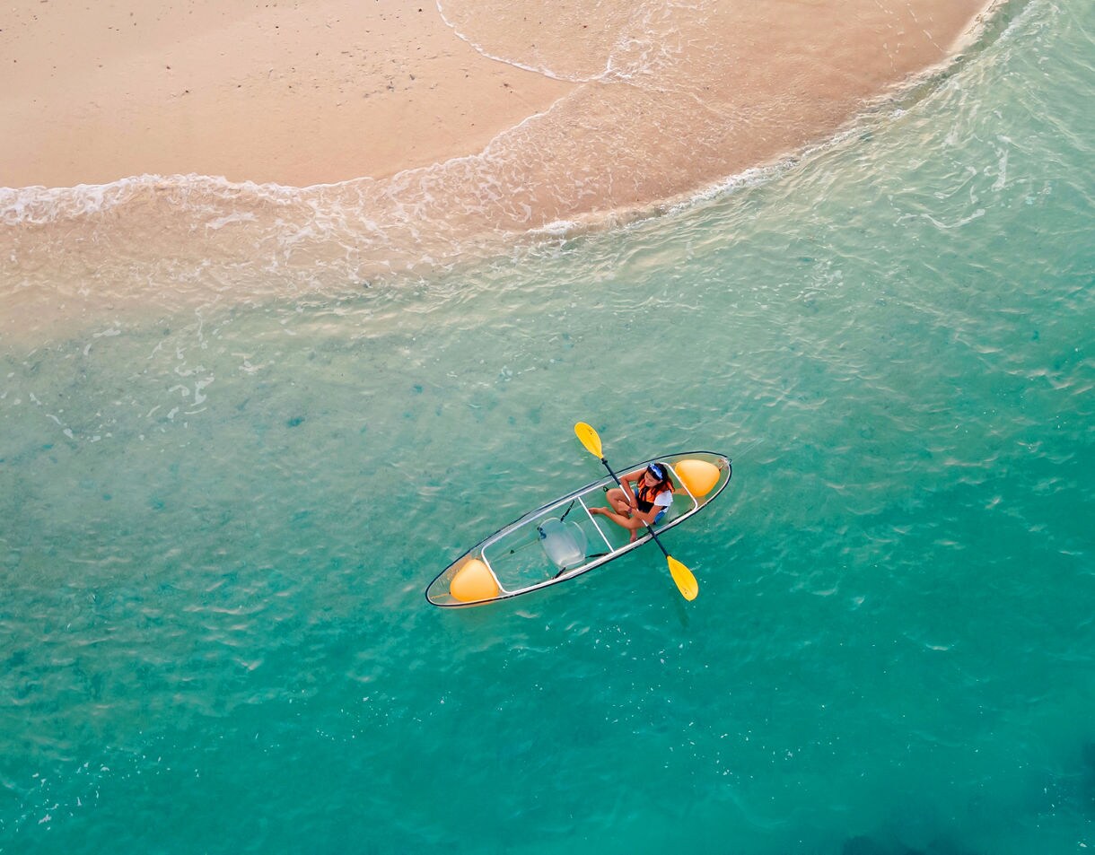 Aerial view of a person paddling a transparent kayak in shallow turquoise water near a pale sandy beach.