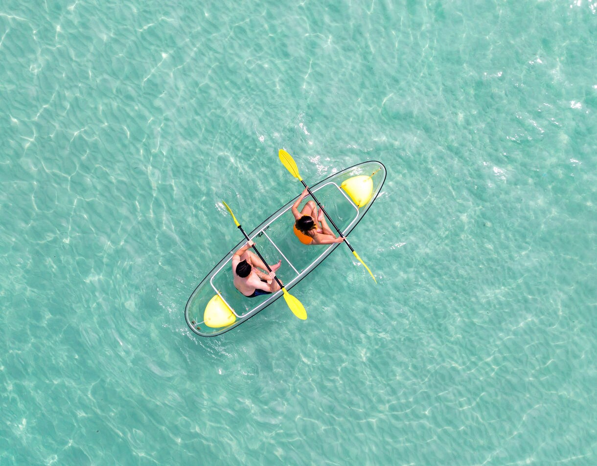 Aerial view of two people paddling a transparent tandem kayak over shallow pale turquoise water with sunlight shimmering on the surface.