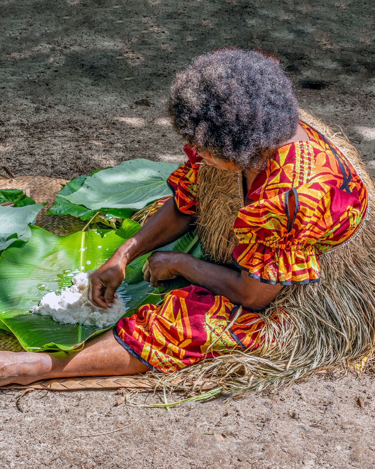 Woman in a bright red and yellow patterned dress kneeling on woven mats while preparing white manioc on large green leaves outdoors.